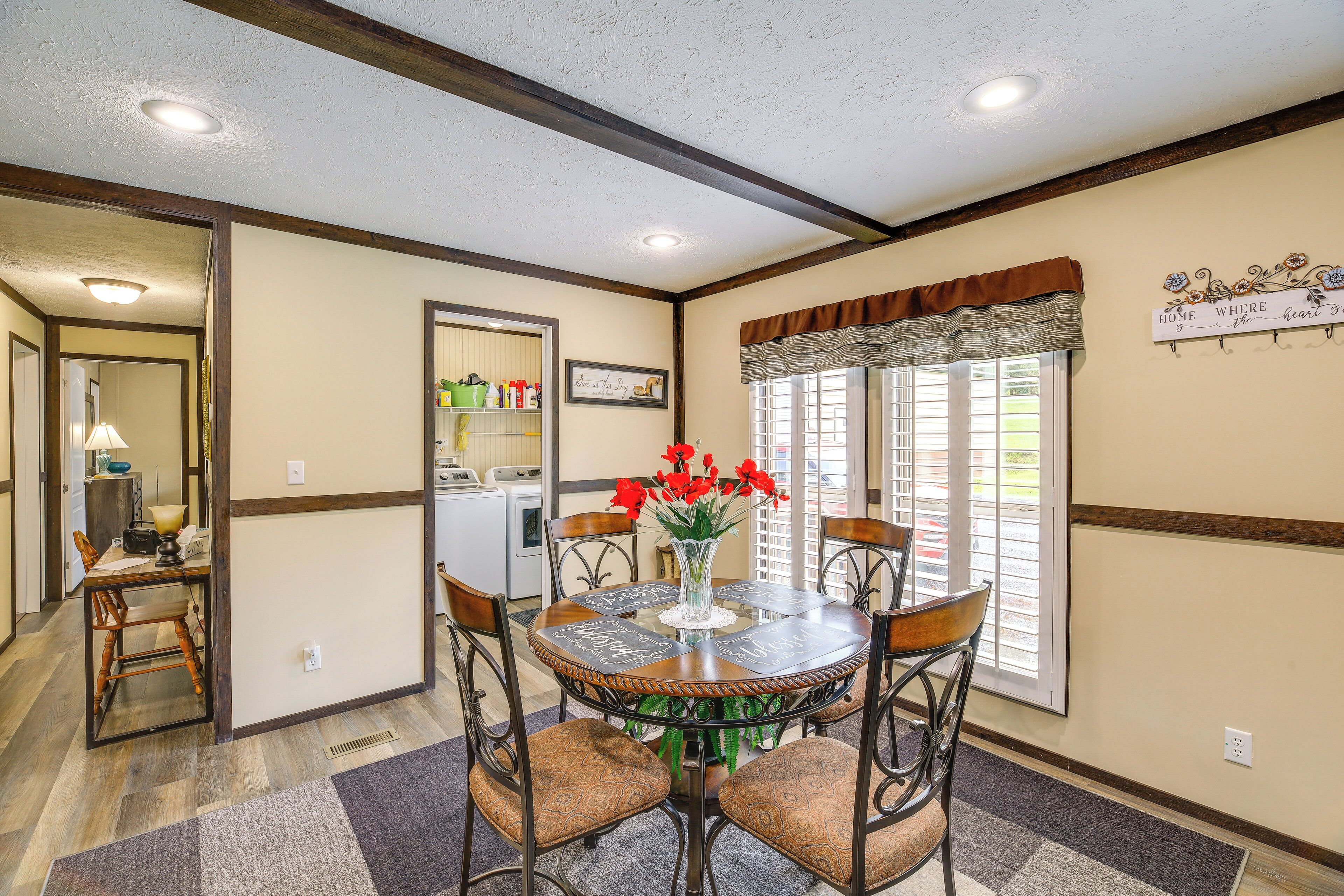 A dining area with a round table, chairs, and a vase with red flowers. There is a window with blinds and a sign on the wall.