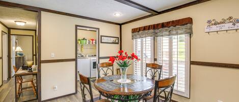 A dining area with a round table, chairs, and a vase with red flowers. There is a window with blinds and a sign on the wall.