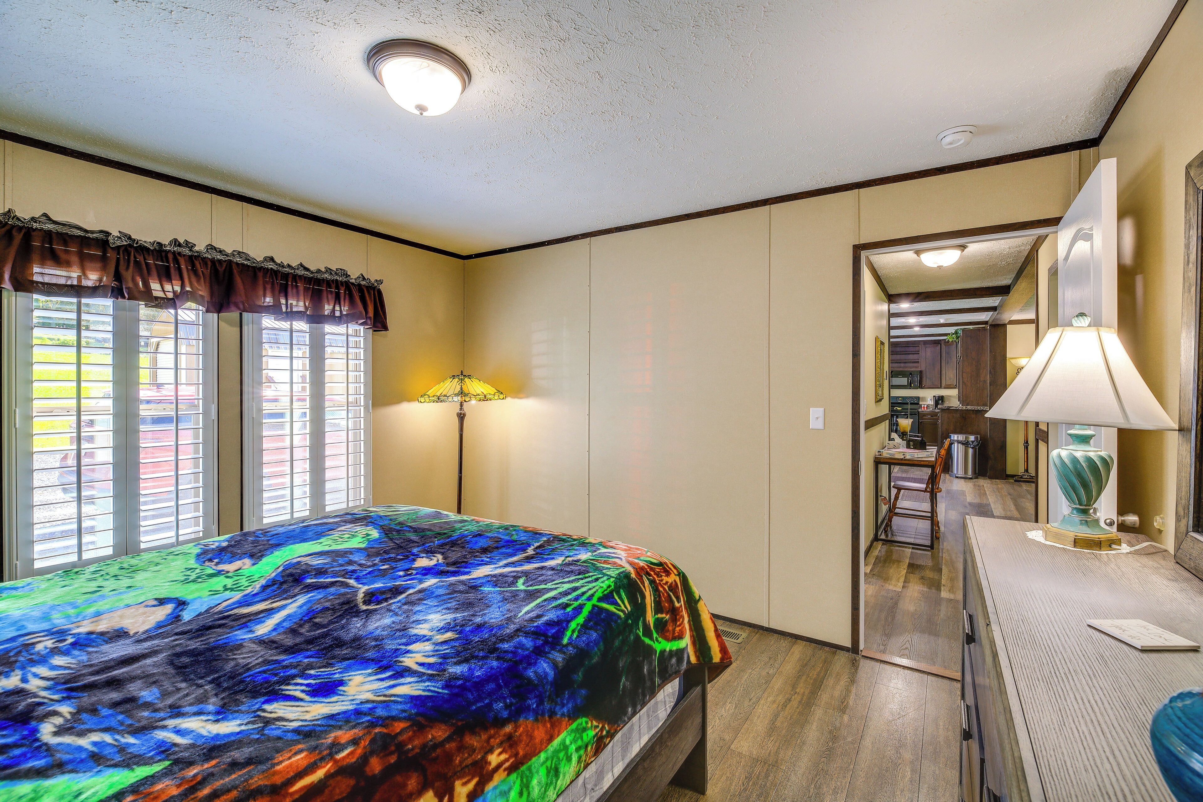 A bedroom with a bed, a desk, a lamp, and a view of a kitchen and hallway.