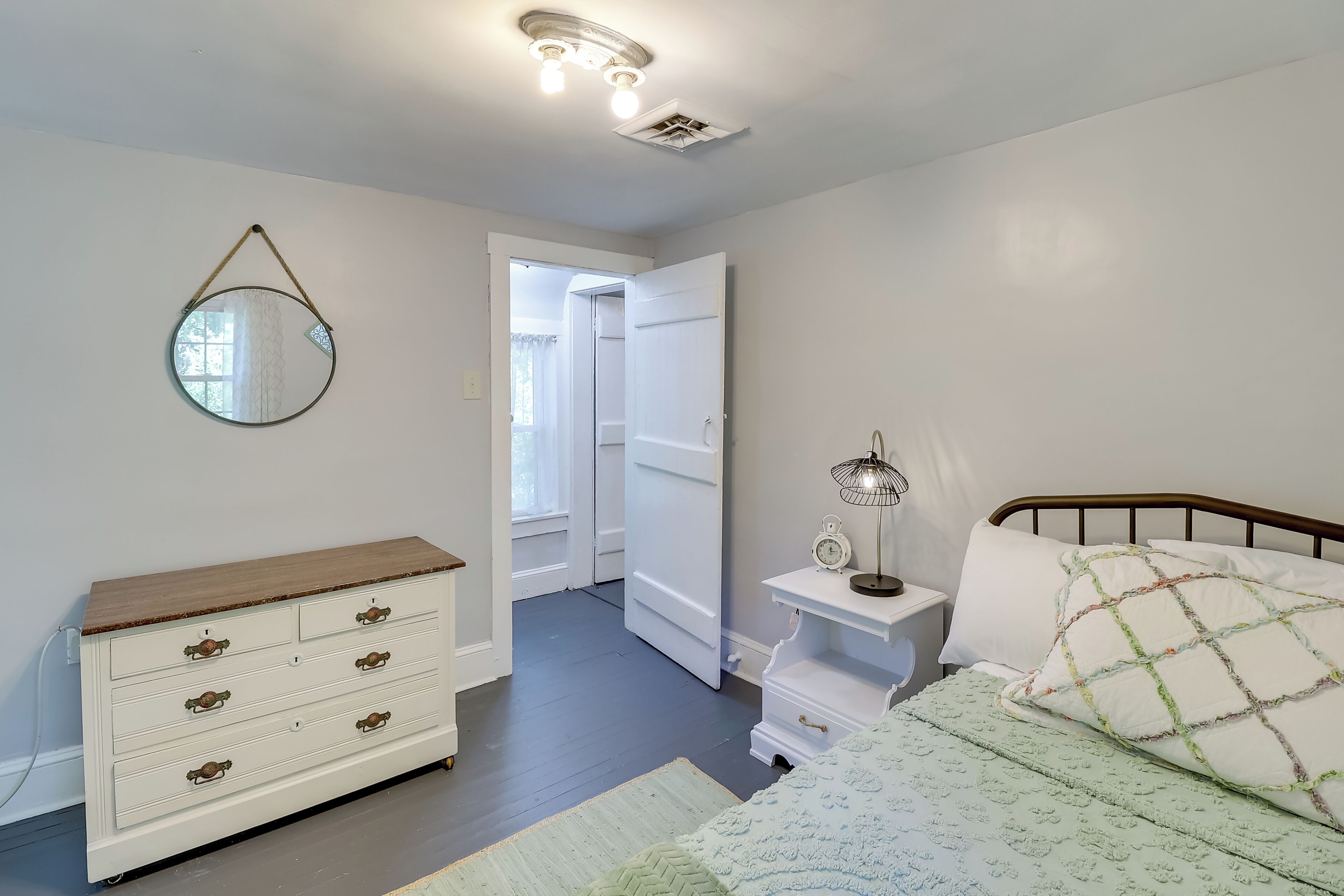 A bedroom with a bed, bedside table, lamp, dresser, mirror, and a door leading to another room.