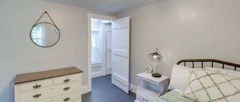 A bedroom with a bed, bedside table, lamp, dresser, mirror, and a door leading to another room.