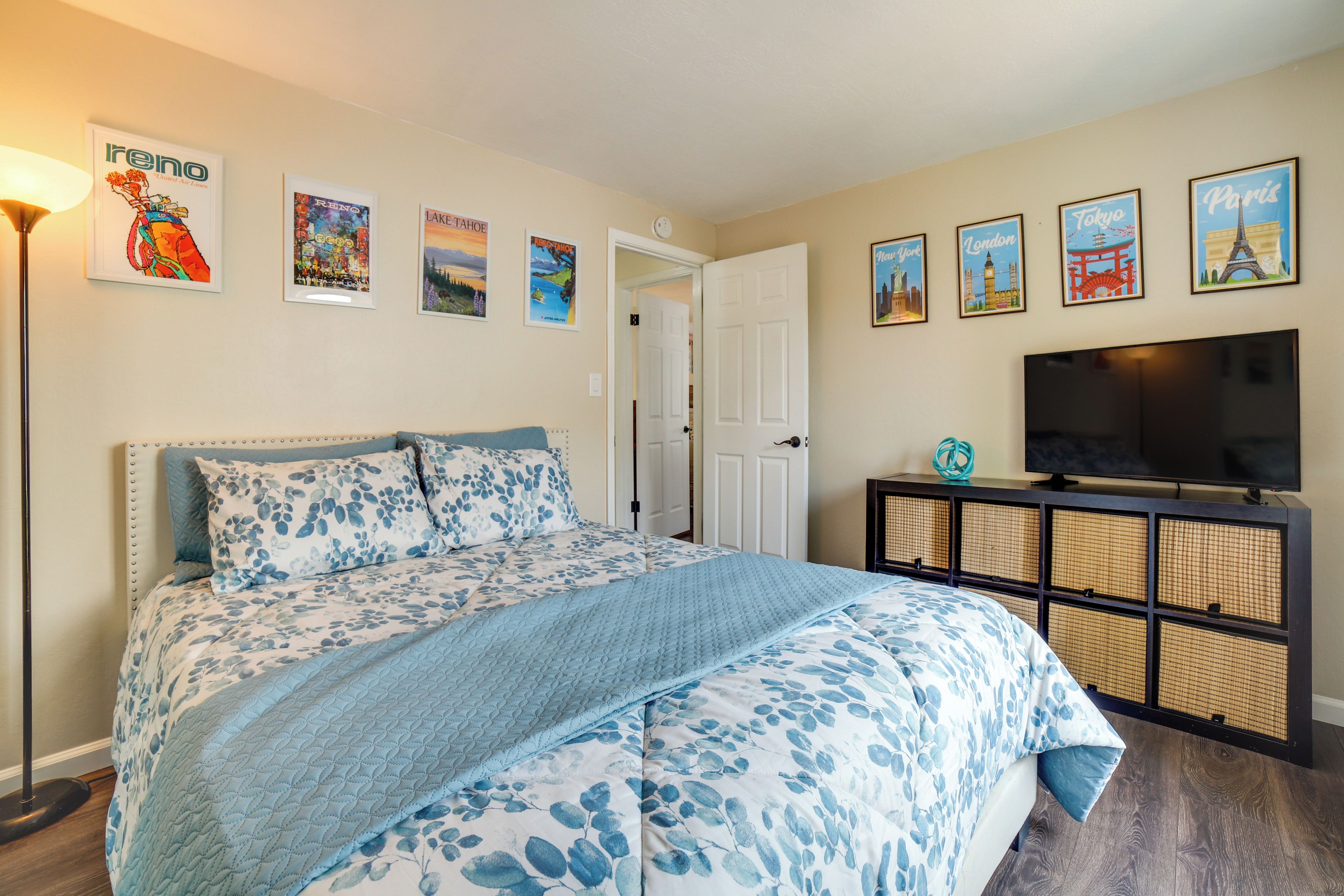 A bedroom with a large bed, a television on a stand, and wall art depicting various cities.