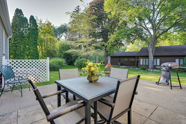 Um pátio externo com mesa e cadeiras, churrasqueira e um jardim com árvores e cerca.