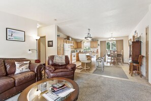 A spacious living room with a brown leather sofa, a coffee table with books and a remote, and a kitchen in the background. - Fort Smith Vacation Rental Near Bighorn River (Fort Smith)