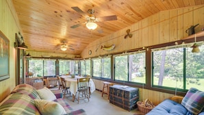 A cabin interior with a dining area, two sofas, a ceiling fan, and large windows. - Grill & Fire Pit: Waterfront Cameron Retreat! (Cameron)
