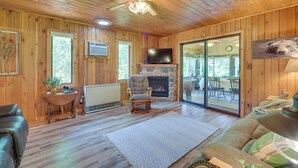 A wooden cabin interior with a fireplace, a ceiling fan, a television, and a large window. - Grill & Fire Pit: Waterfront Cameron Retreat! (Cameron)