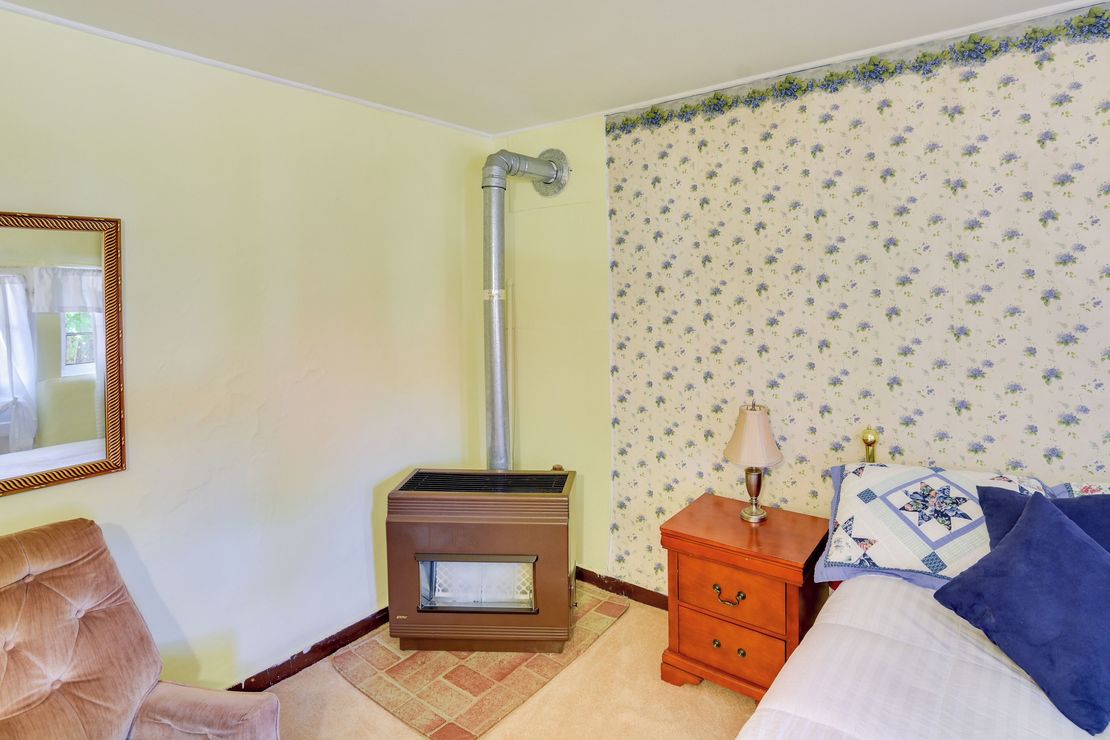 Een kamer met een houtkachel, een bed met blauw-witte beddengoed, een nachtkastje met een lamp en een stoel.