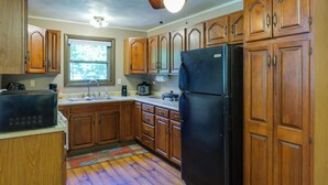 A kitchen with wooden cabinets, a black refrigerator, a window, and a ceiling fan. - Riverfront Covington Cabin w/ Deck & Fire Pit! (Covington)