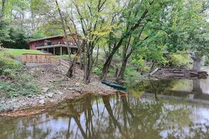 A lakeside cabin with a deck and outdoor seating, surrounded by trees and a calm water body. - Riverfront Covington Cabin w/ Deck & Fire Pit! (Covington)