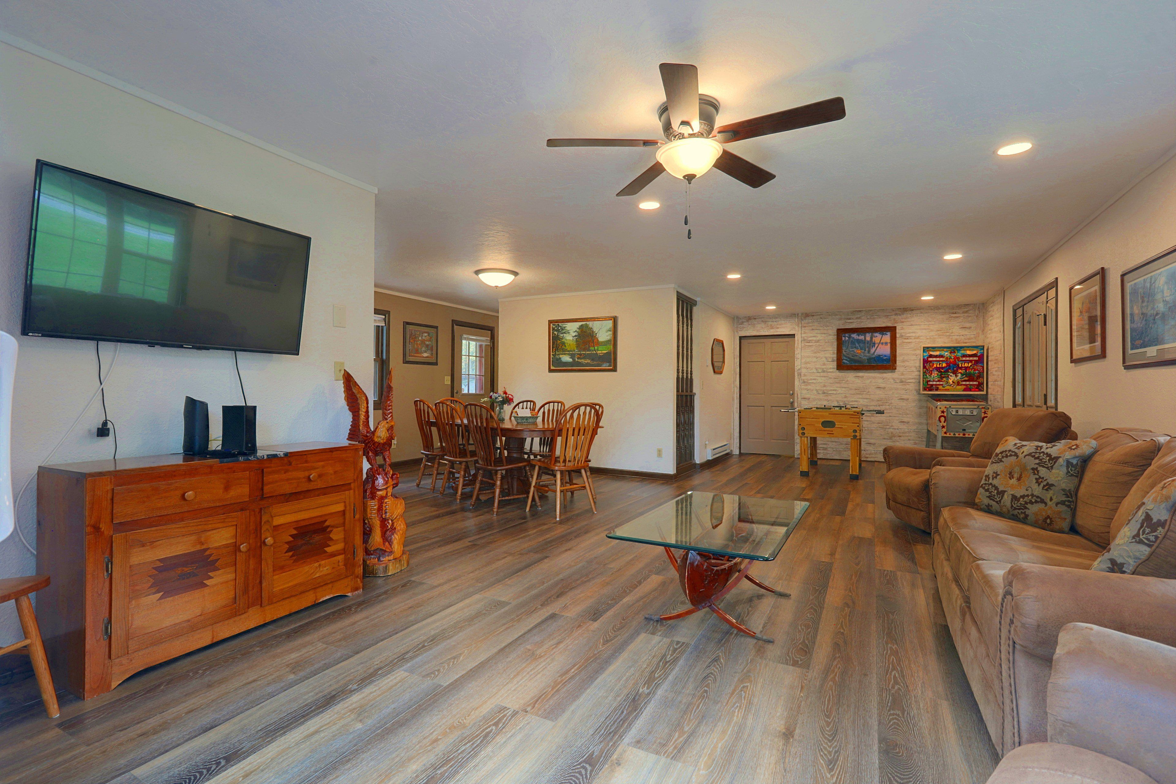 A spacious living room with a flat-screen TV, wooden furniture, a glass coffee table, and a ceiling fan.