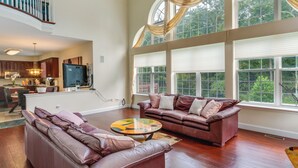 A spacious living room with a large window, a leather sofa, a coffee table, and a kitchen in the background. - Centrally Located Family Home w/ Pool (Harleysville)