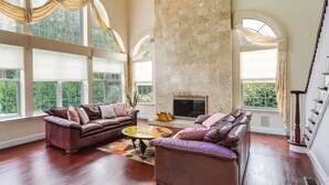 A spacious living room with two leather sofas, a fireplace, and large windows offering a view of greenery. - Centrally Located Family Home w/ Pool (Harleysville)