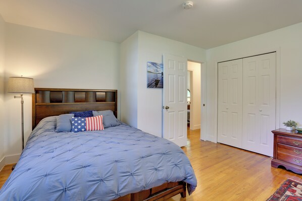 A bedroom with a large bed, a nightstand, a lamp, a dresser, and a door leading to another room.