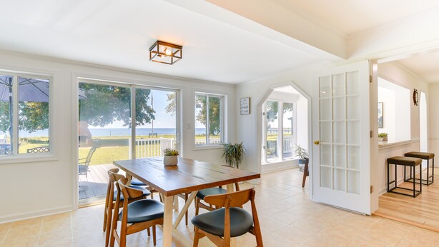 A spacious dining area with a wooden table and chairs, large windows offering a view of the outdoors, and a white door leading to another room. - Sun-dappled Waterfront Home on Potomac River! (Scotland)