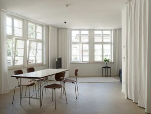 Une pièce spacieuse avec une longue table et six chaises, de grandes fenêtres et une petite table d’appoint sur laquelle est posée une plante.