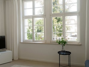 Een kamer met een groot raam, een flatscreentelevisie op een witte kast, en een kleine tafel met een bloemenvaas.