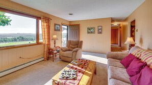A cozy living room with a view of the countryside, a comfortable armchair, a sofa with cushions, and a coffee table with books and a board game. - Muncy Vacation Rental w/ Deck & Views (Muncy)