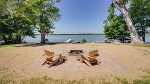 Een terras aan het strand met Adirondack-stoelen en een vuurkuil, met uitzicht op een meer met een pier en boot.