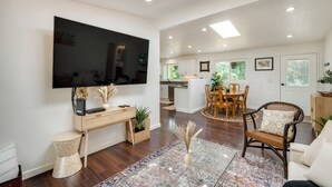 A spacious living room with a large flat-screen TV, wooden flooring, a dining area with a round table, and a kitchen in the background. - Modern Meadow Vista Vacation Home w/ Spacious Deck (Meadow Vista)