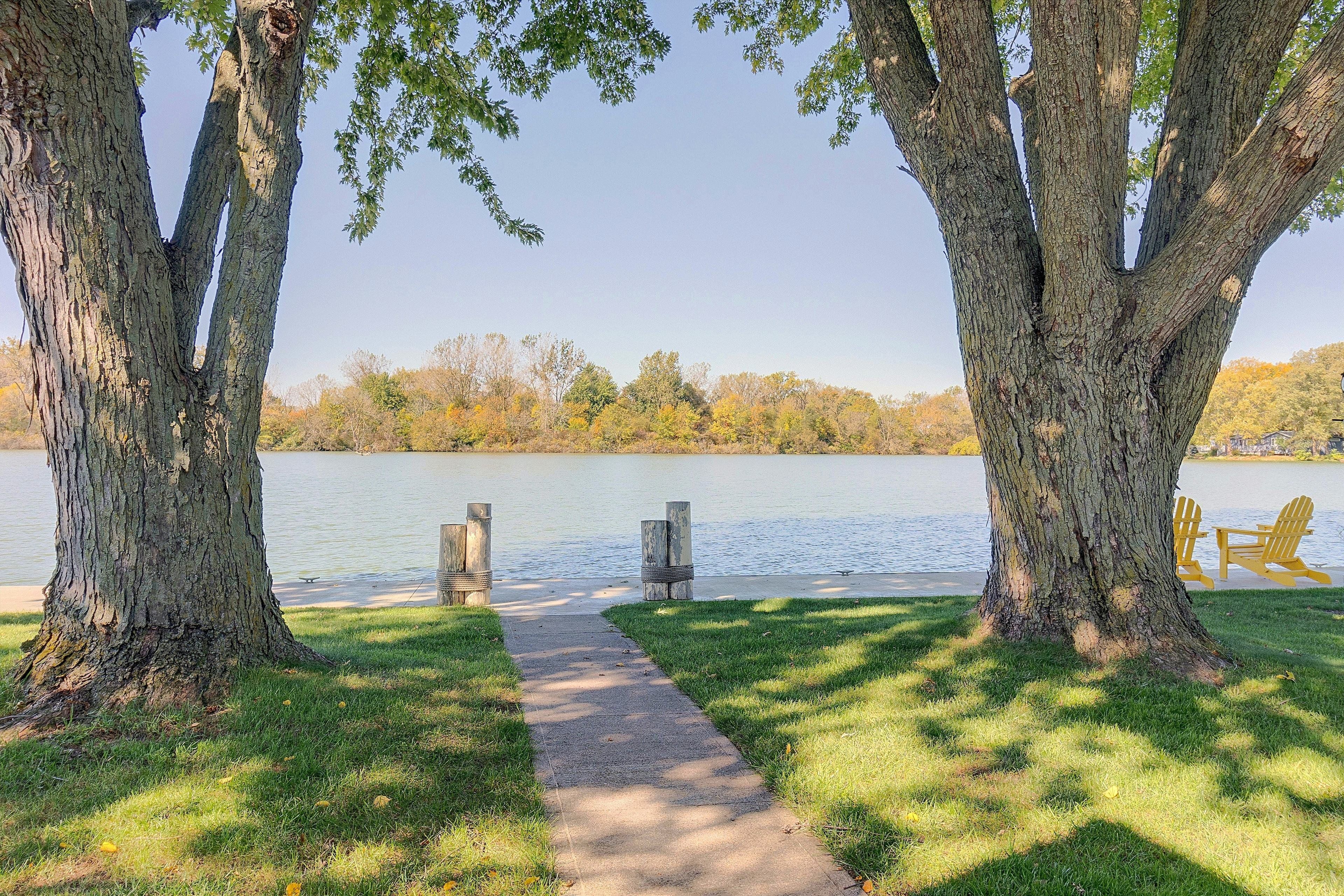 A pathway leading to a lake with trees on either side and a row of Adirondack chairs.