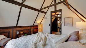 Een slaapkamer met een schuine kap, houten balken, een bed met witte beddengoed, een nachtkastje, een lamp en een boekenkast.