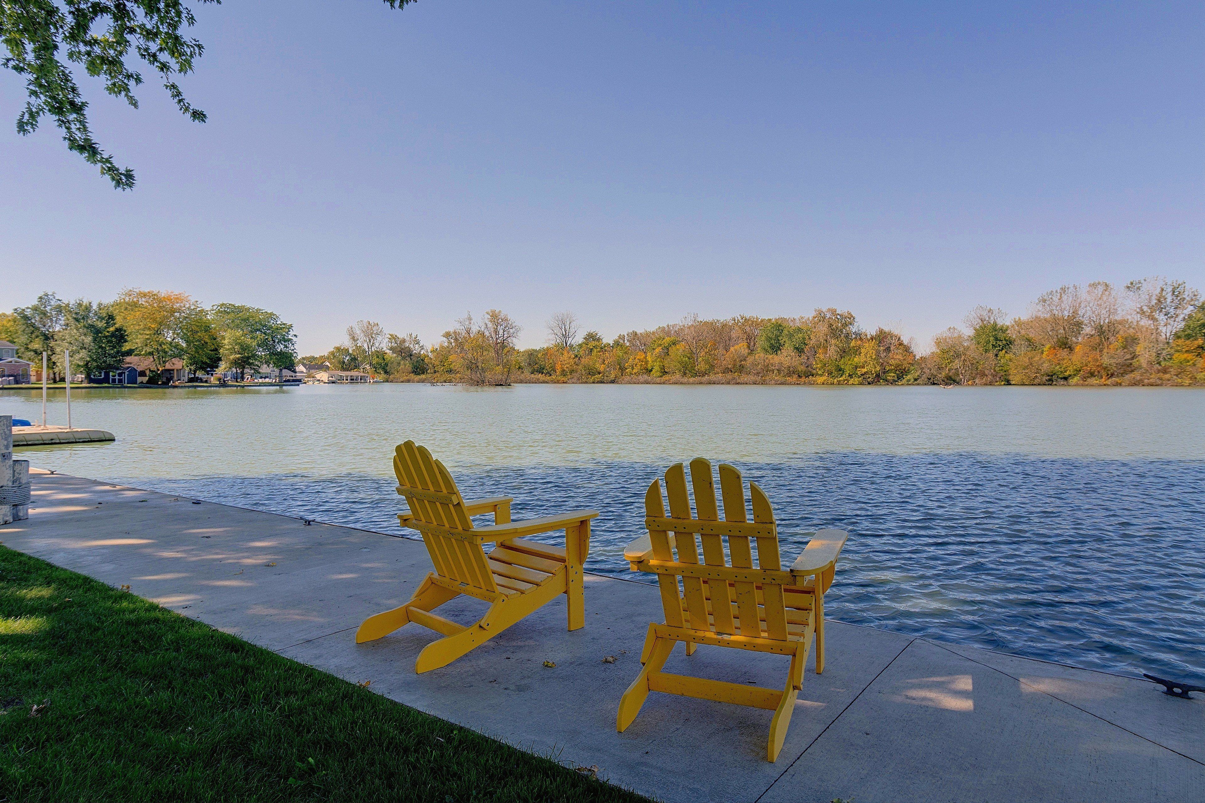 Two Adirondack chairs facing a calm lake with a dock and houses in the background.