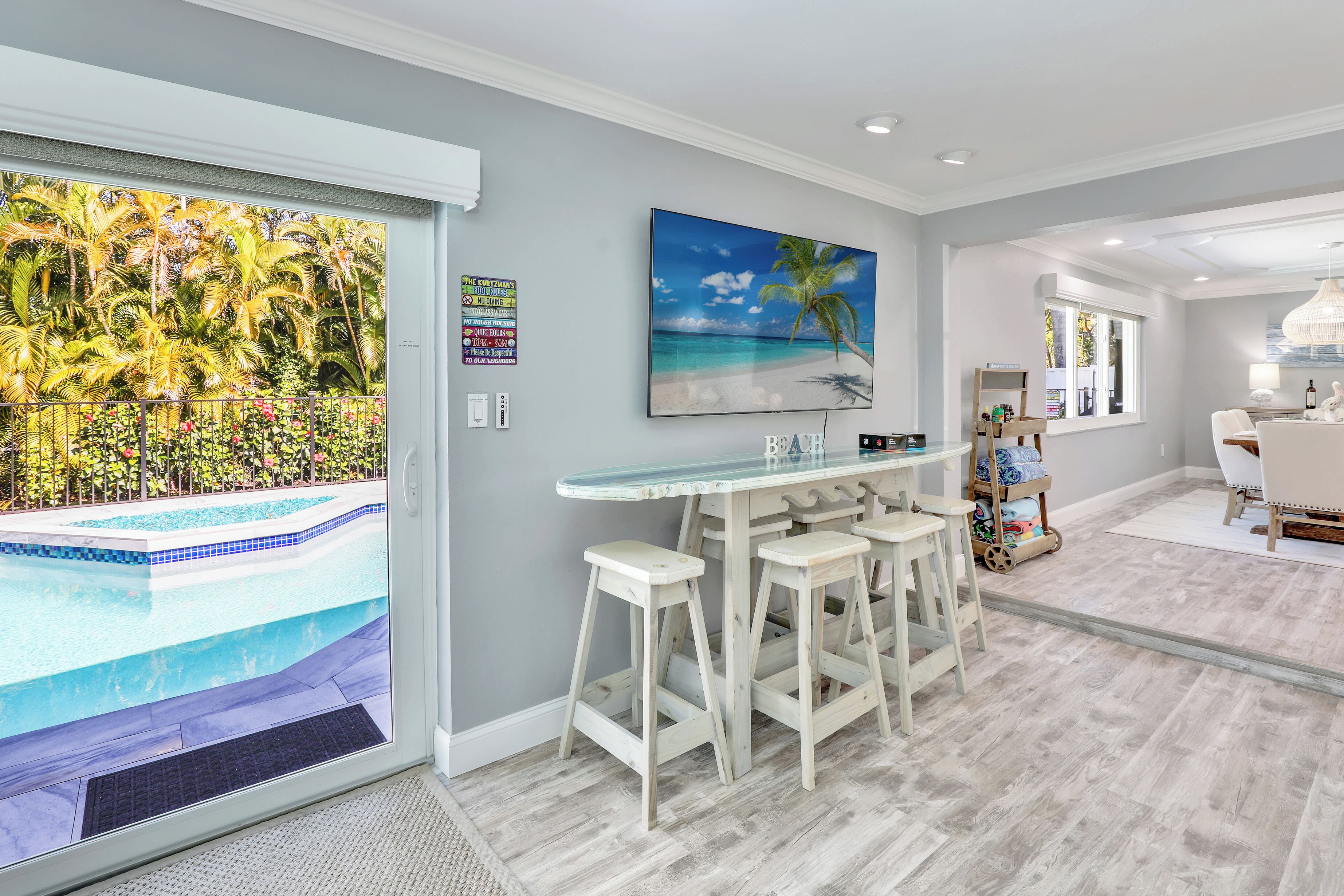 A modern kitchen with a bar area, a large window with a view of a pool, and a tropical-themed painting.
