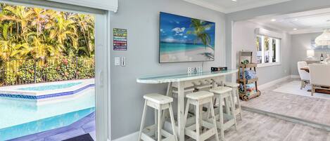 A modern kitchen with a bar area, a large window with a view of a pool, and a tropical-themed painting.