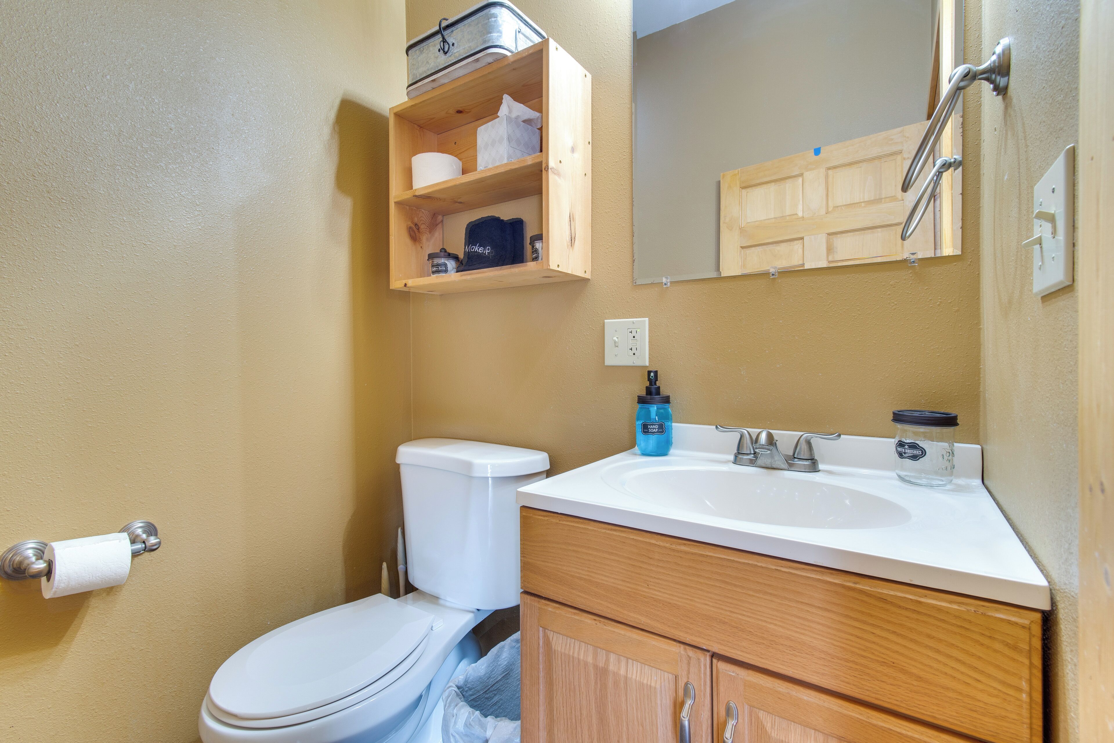 A compact bathroom with a toilet, a sink, wooden cabinets, a mirror, and a small shelf with toiletries.
