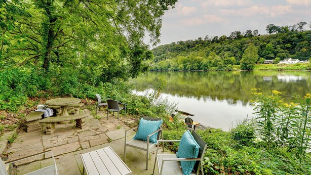 An outdoor patio with stone seating, metal chairs with blue cushions, and a view of a lake surrounded by greenery. - Upper Black Eddy Home w/ Delaware River Access! (Upper Black Eddy)