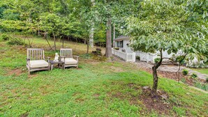 A backyard with two wooden chairs and a small table, surrounded by lush greenery and trees. - Idyllic Cave Spring Studio w/ Patio & Fire Pit! (Roanoke)