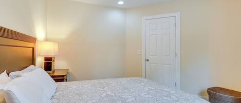 A bedroom with a bed, a nightstand, a lamp, a door, and a bench.