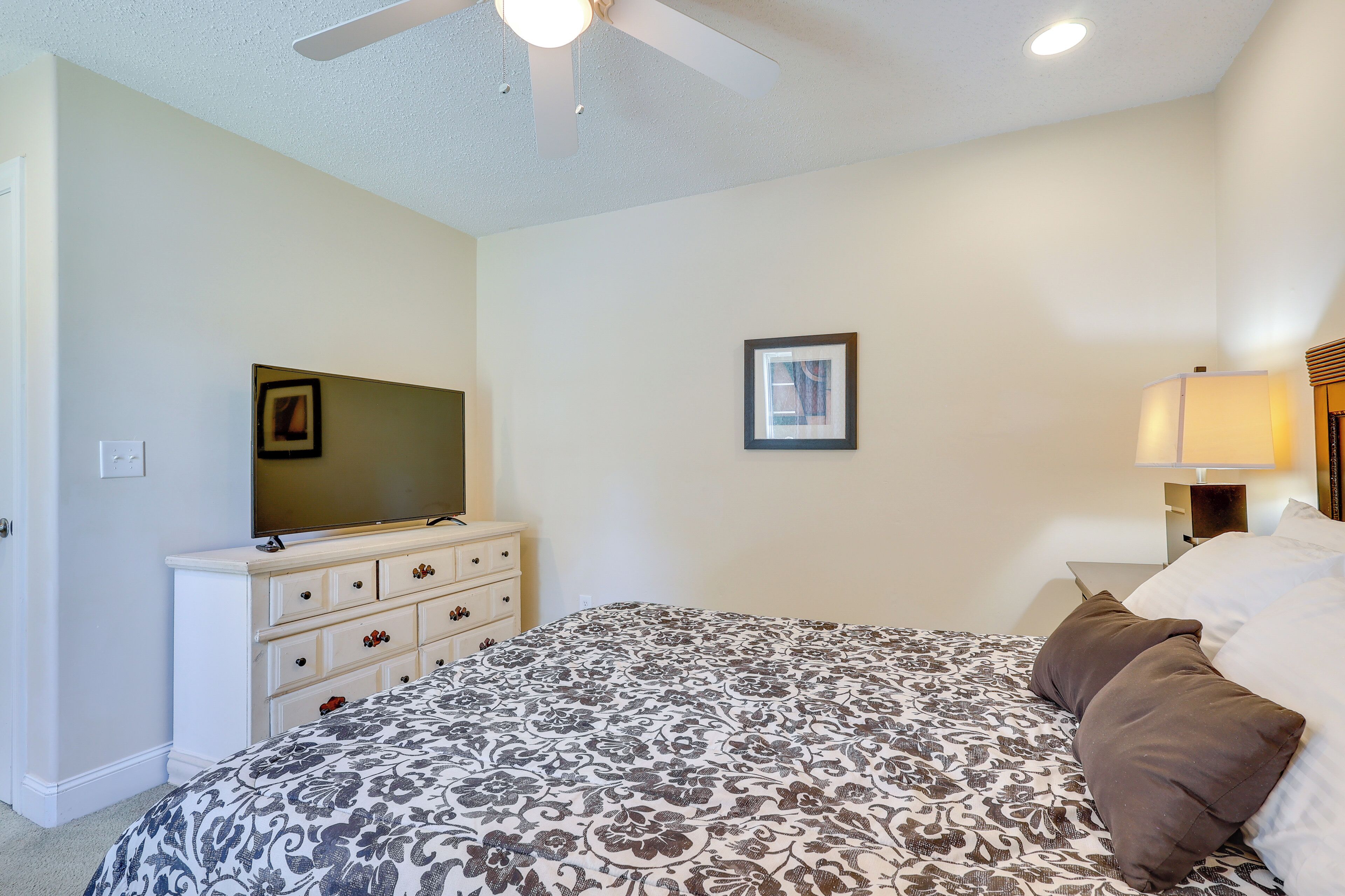 A bedroom with a bed, a dresser, a television, a framed picture, and a nightstand with a lamp.