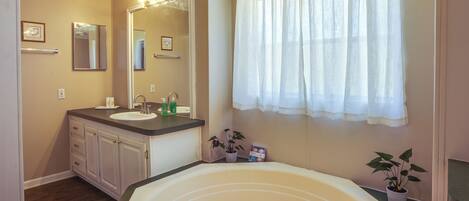 A bathroom with a bathtub, two sinks, a mirror, and a window with a white curtain.
