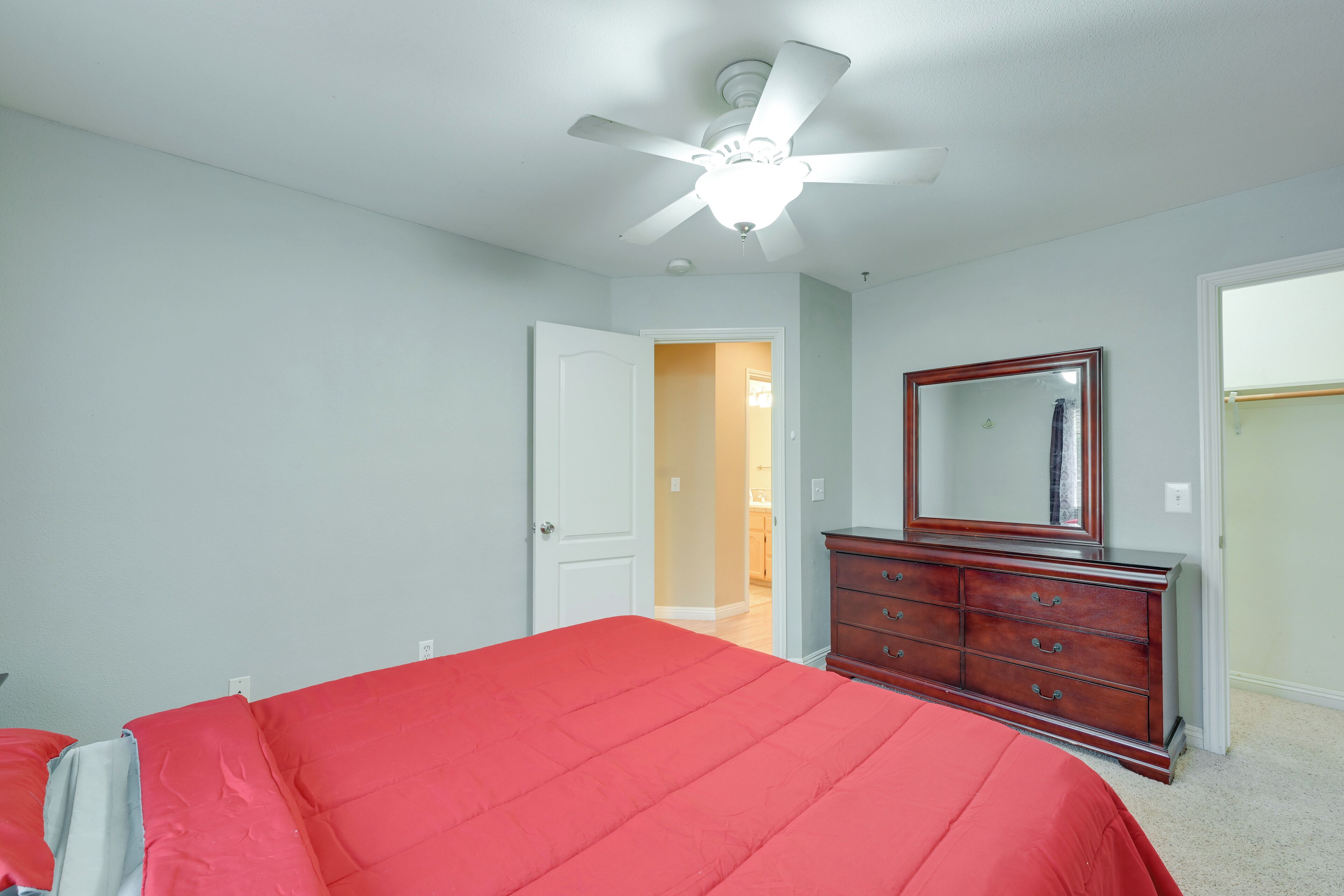 A bedroom with a red bedspread, a wooden dresser, a ceiling fan, and a door leading to another room.