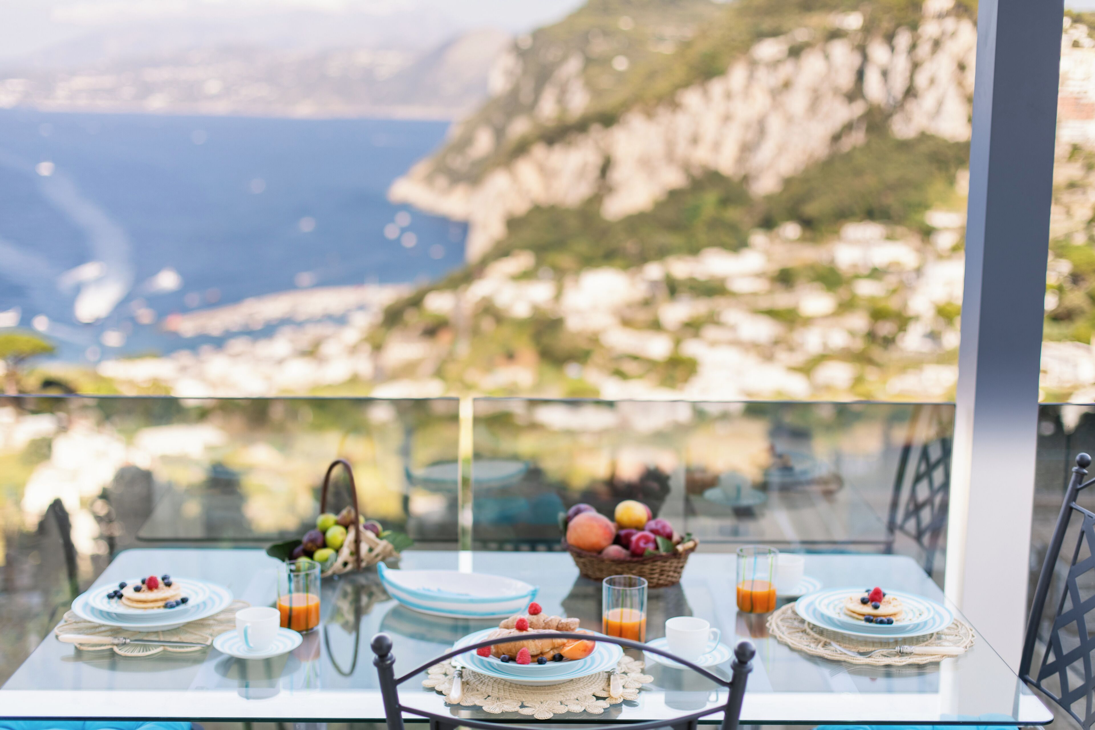 A table set for breakfast with fruit and juice, overlooking a coastal landscape.