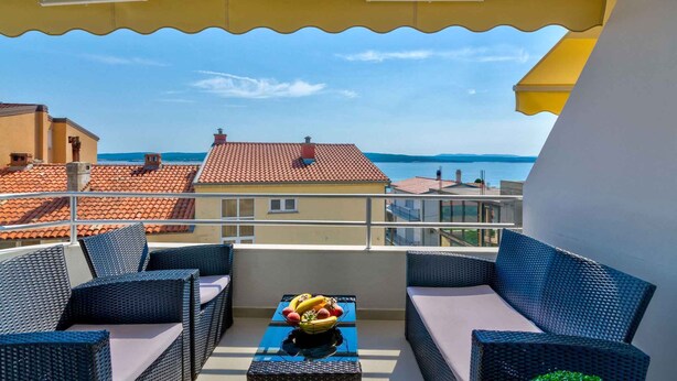 A balcony with wicker furniture and a fruit bowl, overlooking a coastal area with red-roofed buildings. - Holiday apartment mit Meerblick (Crikvenica)