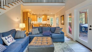 A spacious living room with a blue sectional sofa, a leather ottoman, and a kitchen in the background.