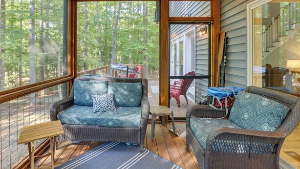 A covered porch with wicker furniture, a glass door leading to a deck, and a view of a forested area.