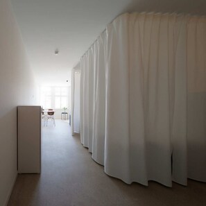 Een ruime, goed verlichte gang met een modern, minimalistisch design. Aan één kant hangt een lang, wit gordijn en aan de andere kant een witte deur, die leidt naar een kamer met een kleine tafel en stoelen.