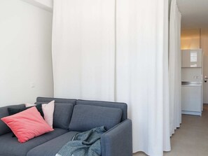 Een moderne woonkamer met een grijze bank, witte gordijnen en een kitchenette op de achtergrond.