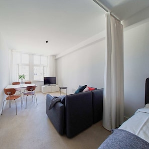 Een moderne, goed verlichte kamer met een donkere bank, een eettafeltje met stoelen, een televisie en een groot raam met gordijnen.
