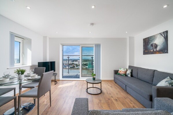 A modern living room with a glass dining table, grey sofas, and a city view through large windows.