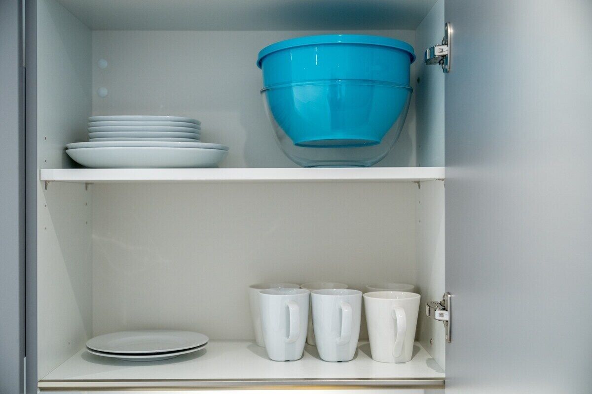 Un meuble de cuisine avec des étagères blanches où sont rangées des assiettes, un saladier bleu et des tasses blanches.