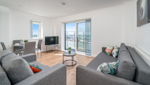A modern living room with a grey sofa, a TV, a dining table with chairs, and a large window offering a city view.