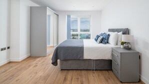 A modern bedroom with a large bed, a grey dresser, and a city view through a large window.