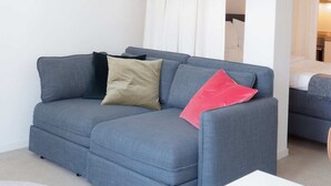 Un salon moderne avec un canapé gris, une table basse blanche et une vue sur une chambre à coucher à travers un rideau blanc.