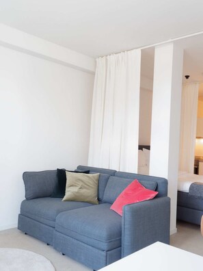 Een moderne woonkamer met een grijze bank, een witte salontafel en een zicht op een slaapkamer door een witte gordijn.