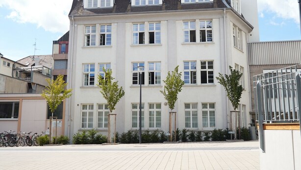 Un immeuble à plusieurs étages, doté d’un toit plat, de grandes fenêtres et d’un espace extérieur pavé agrémenté d’arbres et d’un support à vélos.
