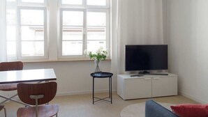 Un salon moderne doté d’un téléviseur à écran plat, d’une table à manger avec des chaises, d’une petite table d’appoint sur laquelle est posé un vase, et d’une fenêtre avec des rideaux.